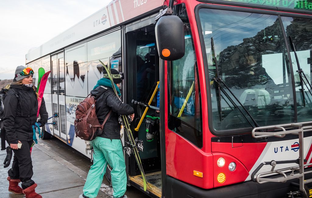 People boarding a red uta bus at a ski resort, carrying ski gear, dressed in winter clothing. a snowy mountain landscape is visible in the background.