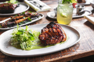 A grilled chicken breast with a side of green salad, served on a white plate with a wooden table background. other dishes and a glass of iced drink are also visible.