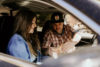 A man in a plaid shirt and cap hands a sandwich to a woman in a denim jacket, both seated in a car with the window down.