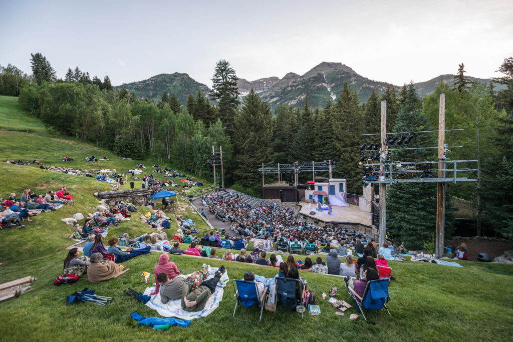 An outdoor concert with people seated on grass and bleachers, enjoying a performance in a natural amphitheater surrounded by lush green mountains under a twilight sky.