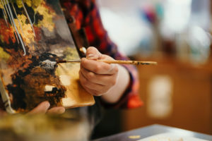 Close-up of a painter's hand holding a brush against a colorful palette, focusing on applying paint to a canvas in a warmly lit studio.