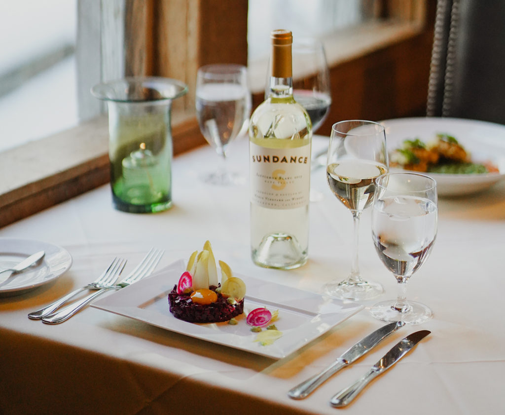Elegant dining setup by a window with natural light, featuring a plate of gourmet sundance food, two glasses of white wine, and a bottle labeled "sundance".