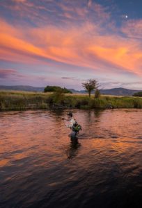 A person wearing fishing gear stands in a river casting a fishing line at sunset. The sky is illuminated with vibrant orange and pink hues as the sun sets. Trees and distant mountains are visible in the background. A small crescent moon appears in the sky.