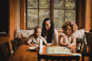 A mother and her two children, a boy and a girl, are sitting at a wooden dining table playing a board game in a cozy room with large windows showcasing a lush green background.