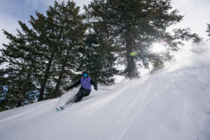 A skier in a purple jacket glides gracefully down the mountain with the sun behind them.