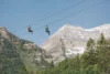 Two people zip-lining against a scenic backdrop of a snowy mountain peak and lush green forests under a clear blue sky.