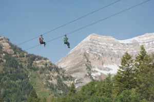 Two people zip-lining against a scenic backdrop of a snowy mountain peak and lush green forests under a clear blue sky.