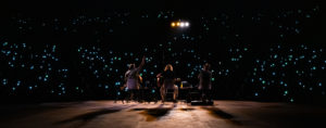 Night-time concert with three musicians and a crowd full of people holding up lights.