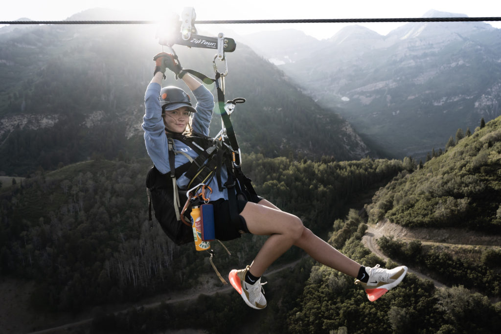 A woman ziplining over a lush mountain valley, smiling at the camera, equipped with a safety harness and helmet, during a sunny day.