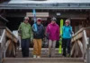 Four cheerful skiers in colorful winter gear walking down wooden stairs together, holding skis and poles, with a ski resort building in the background.