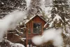 A cozy wooden cabin surrounded by snow-covered pine trees, viewed through a blurred foreground of snowflakes, in a wintry mountainous landscape.