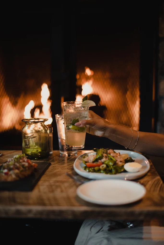 A cozy dining scene featuring a person's arm reaching for a glass of iced drink beside a plate of salad, with a glowing fireplace in the background, conveying a warm, intimate ambiance.