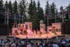A vibrant outdoor theater performance is taking place on a well-decorated stage resembling a Mediterranean village. Actors dressed in colorful costumes are animatedly performing under the evening sky, surrounded by tall trees and an engaged audience.
