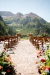 An outdoor wedding venue with wooden chairs arranged in rows facing a mountainous backdrop. Colorful flower arrangements line the aisle and frame the view of lush, green mountains under a clear blue sky.