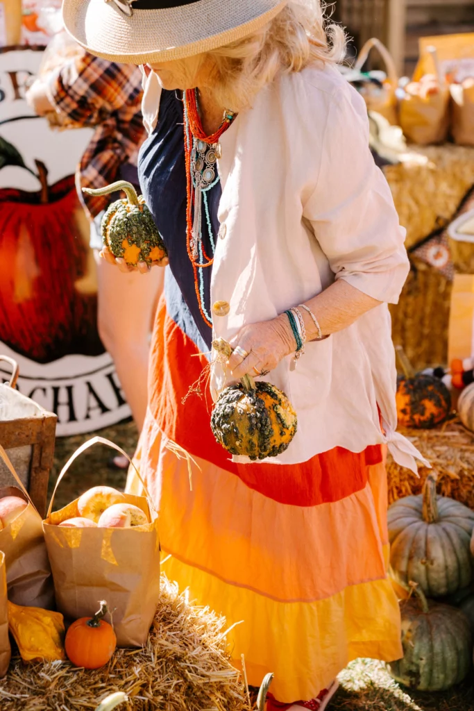 A woman in a colorful skirt and hat holds decorative gourds at the Sundance Harvest Market, surrounded by pumpkins and autumnal decorations.