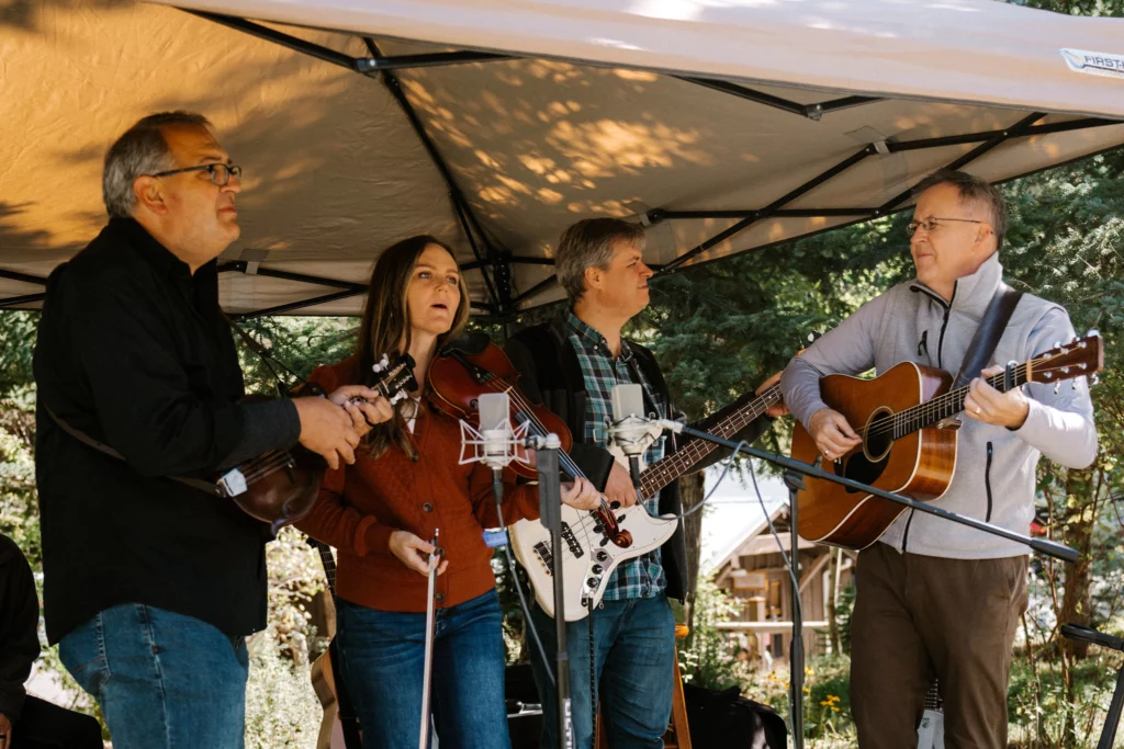Four musicians perform at the Sundance Harvest Market outdoors under a canopy, each playing different instruments: a violin, a mandolin, an electric bass, and an acoustic guitar.