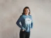 A joyful woman wearing a blue long-sleeve shirt with a graphic print stands with one hand on her hip, smiling against a neutral-toned background.