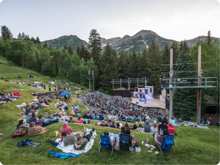 Outdoor concert with attendees sitting on grass and in seats at a venue surrounded by mountains. people are on blankets and folding chairs, enjoying the event in a scenic natural amphitheater.