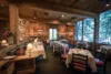 Interior of a cozy, rustic-themed restaurant with colorful table settings, wooden walls and ceiling, subtle lighting, and windows offering a view of greenery outside.