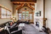 A cozy, well-lit living room featuring a stone fireplace, wooden ceiling beams, leather sofas, red armchairs, and a large coffee table, with windows covered by wooden blinds.