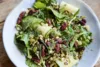 A fresh salad featuring zucchini ribbons, mixed greens, pecans, pumpkin seeds, and cranberries garnished with dill, served in a white bowl on a wooden table.