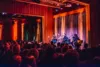 Three musicians perform on stage in a warmly lit auditorium, playing acoustic guitars to an audience of attentive spectators. the setting features wooden paneling and purple curtains.