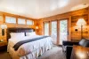 Cozy, wood-paneled bedroom featuring a king-sized bed with white and grey bedding, a leather armchair, a bedside lamp, and glass doors leading to a balcony.