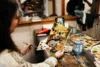 A woman in a cozy art studio paints a colorful landscape on a small canvas, surrounded by brushes and palettes.