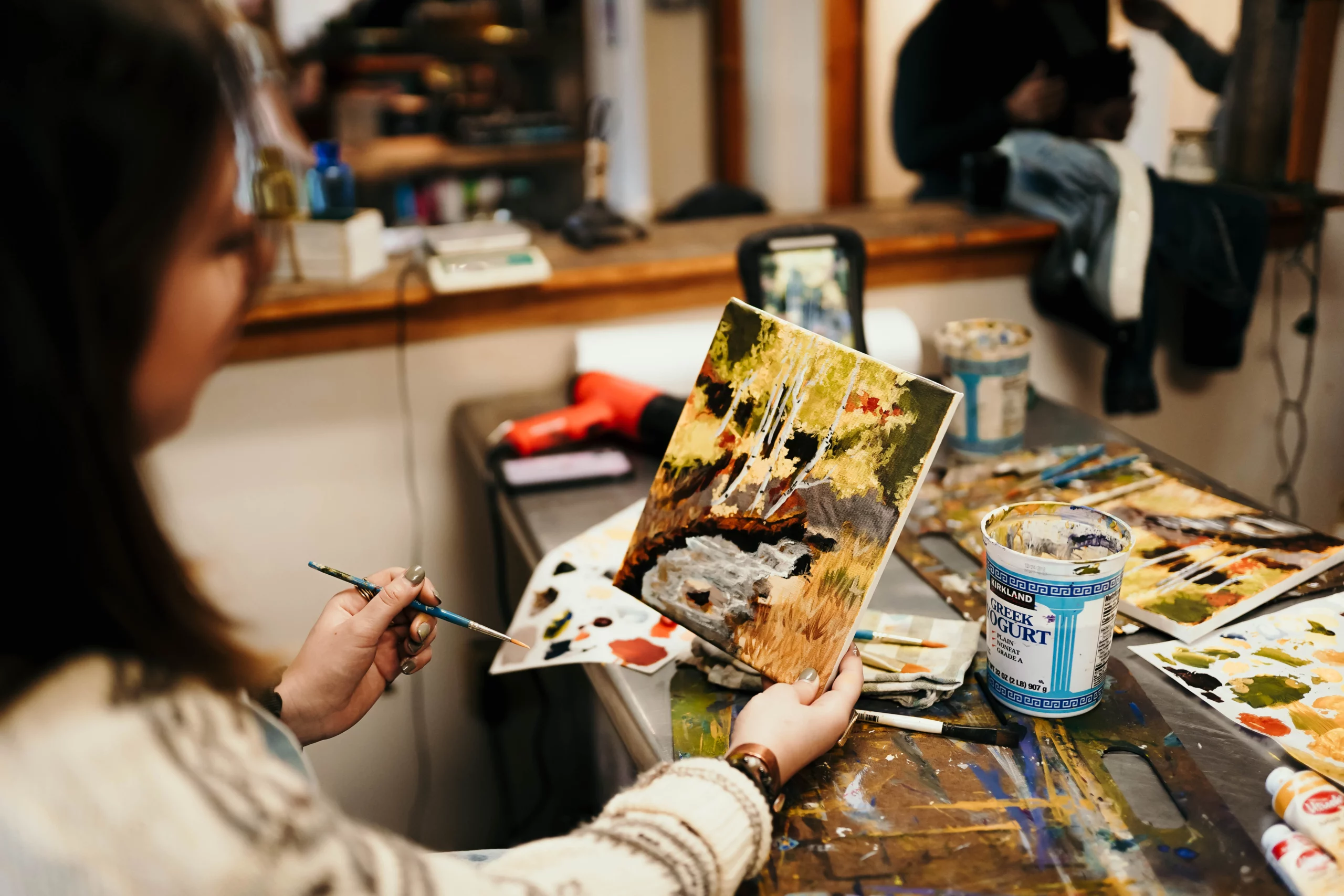 A woman in a cozy art studio paints a colorful landscape on a small canvas, surrounded by brushes and palettes.