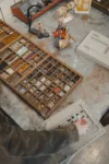 A person works at a cluttered workshop table, organizing assorted small parts and tools into a compartmentalized tray.