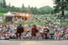 A view from behind musicians on an outdoor stage facing a large audience gathered in a natural amphitheater surrounded by trees and hills at dusk.