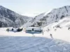 A scenic winter view showing skiers and snow-covered buildings on a mountainside with a frozen lake visible in the valley below under a bright sunny sky.