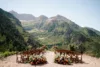 Outdoor wedding setup with rows of wooden chairs and floral decorations, facing majestic green mountains under a clear blue sky.
