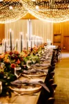 A long banquet table elegantly set for an event, adorned with vibrant floral centerpieces, tall candles, and gold chargers under ambient string lights.