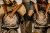 Two people in aprons using pottery wheels to shape wet clay into bowls, focused on their craft amidst a workshop setting with clay splashes and tools visible.