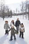 A family of five, including three kids and two adults, smiling and holding hands while standing on a snow-covered path in a wintery, tree-lined landscape. they are all wearing winter jackets and snow boots.