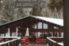 A cozy cabin with large windows, surrounded by snow-covered trees, featuring a terrace with wooden railings and red chairs. a faded sign hangs in the foreground.