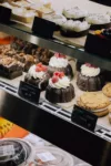 A display case filled with a variety of desserts, including chocolate bundt cakes topped with raspberries and cream, doughnuts, and cookies, all neatly labeled with prices.