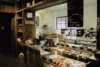 Interior of a cozy deli cafe with a variety of pastries displayed in a glass case, a chalkboard menu hanging above, and coffee-making equipment on the counter.