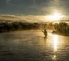 A person in a hat stands fishing in a shimmering river at sunrise, with golden light filtering through mist and surrounding foliage.