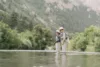 Two people are standing in a river surrounded by forested mountains, fishing. they are dressed in outdoor gear and hats, with one person guiding the other in casting a fishing rod.