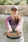 A joyful woman wearing a cap, sunglasses, and outdoor attire, holding a brown trout with both hands, standing in a river. she is smiling, showcasing her catch proudly.