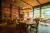 A rustic dining room with wooden walls adorned with various artwork and tools, featuring a large wooden table with chairs, a chandelier overhead, and large windows overlooking greenery.