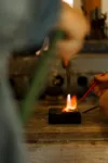 A jeweler uses a torch to heat a metal piece on an anvil, focusing intently on the task, with tools and workshop elements slightly blurred in the foreground and background.