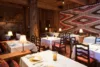 Cozy rustic restaurant interior featuring wooden walls and beams, white tablecloths on tables with elegant place settings, small lamps, and a large patterned wall hanging.