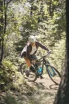 A mountain biker in a red plaid shirt and helmet rides aggressively down a forest trail, surrounded by lush green trees and dappled sunlight.