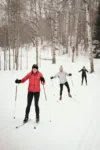 Three people cross-country skiing on a snow-covered trail amongst birch trees; the person in front is wearing a red jacket and black helmet.