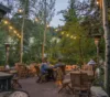 People sitting at wooden tables on a patio surrounded by trees, illuminated by string lights and outdoor heaters, with a view of a forested hill in the background.