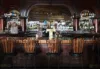 A vintage bar with wooden stools lined up in front, a mirror-backed shelf filled with assorted liquor bottles, and ornate dark wood details.