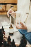 A person dispenses liquid from a dropper into a glass perfume bottle labeled with a large 's' at a workshop table surrounded by other dark bottles.